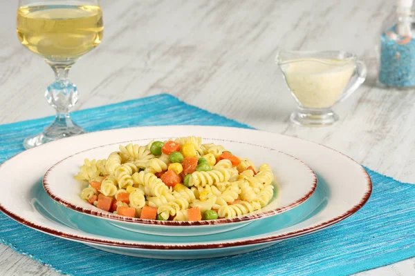 Delicious homemade fusilli pasta cooked with basil pesto, carrots, corn and peas in a plate on a turquoise blue napkin, served with glass of white wine and mustard-mayonnaise sauce