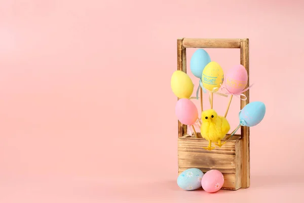 Розовые, желтые и синие окрашенные яйца на палочках и милая курица в деревянной корзине для цветов на розовом фоне. Пасхальная концепция, пространство для текста и дизайна — стоковое фото