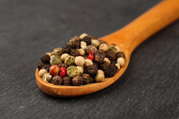 Macro shot di pepe nero, verde, bianco e rosso mescolare in un cucchiaio di legno su una tavola da cucina in pietra scura. Profondità di campo ridotta — Foto Stock