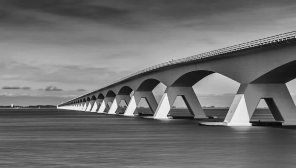 black and white image of bridge over river and cloudy sky