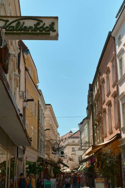 Krems, Avusturya 'da açık mavi gökyüzü altında renkli bina cepheleri, dükkanları, kafeleri ve yayaları olan hareketli bir caddenin fotoğrafı.
