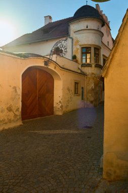 Avusturya 'nın Drnstein kentindeki çok eski bir caddede sarı bir çit, metal armalı kemerli ahşap kapı, güneş ışığında parke taşı yolu ve uzakta beyaz bir ev bulunuyor. Miras ve seyahat projeleri için mükemmel..