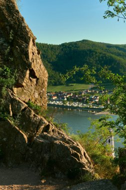 Avusturya 'nın Wachau Vadisi' ndeki Tuna Nehri üzerindeki kaya parçalarından manzaralı tepe manzarası. Kamp alanı karşı kıyıda görünür, arka planda ormanlık tepeler güneşli bir günde.