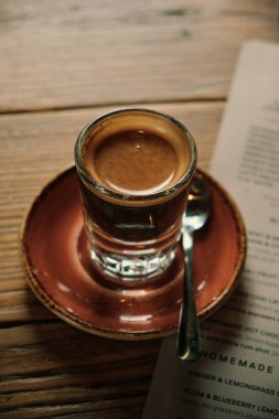 Seramik çay tabağında cam bir fincan kahve, yanında da bir parça menü, ahşap bir yüzeye yerleştirilmiş, rahat ve davetkar bir kafe atmosferi yaratıyor..