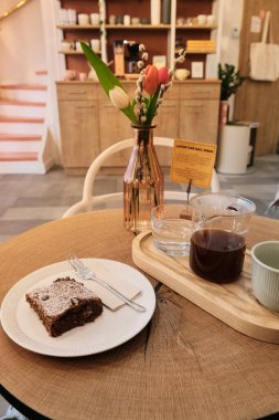 Konforlu kafe sahnesi ahşap masa diliminde, üzerinde çikolatalı browni ve çatal bulunan beyaz tabak, cam sürahi, bardak ve söğüt dallı cam vazo..