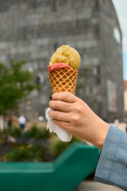 Gri bir binanın arka planında iki kepçeli dondurma külahı ve çilekli bir el, şehir ortamında renkli yaz ziyafetini vurguluyor..