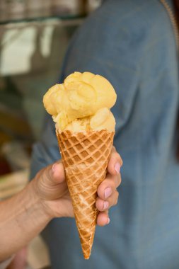 Gri ceketin arka planına karşı mango dondurma külahını tutan bir elin yakın çekimi, canlı yaz ziyafeti ve gelişigüzel şehir tarzını vurguluyor..