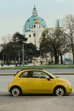 Viyana 'da Karlskirche yakınlarında park edilmiş parlak sarı bir araba. Yeşil kubbe ve sütun görünür, bulutlu gökyüzü, kışın sonu / ilkbahar başlangıcı atmosferi.