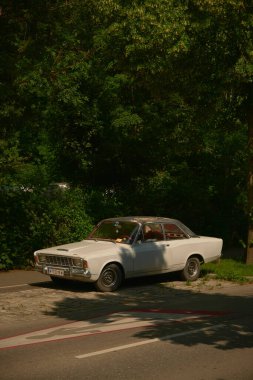 Kırmızı direksiyonlu klasik beyaz araba ve ormanın yakınına park edilmiş kırmızı koltuklar. Ön planda görünen park çizgileri, doğada klasik otomobil sahnesi.