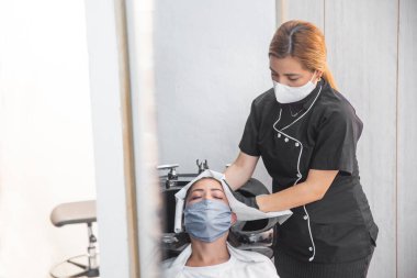 Koruyucu maskeli stilist müşterinin saçını havluyla kurutuyor.