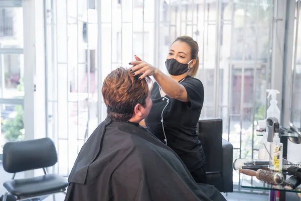 Stylist with protective mask combing hair of a female client at the hair salon