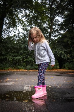 Etrafta sarı sonbahar yaprakları, çocukluk kavramı, sonbahar sezonu ve açık hava oyunu ile su birikintisinin yanındaki ıslak kaldırımda duran pembe çizmeli çocuk.