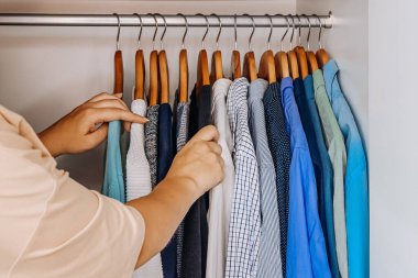 Close-up of female hand touching and choosing shirts on wooden hangers inside a neatly organized wardrobe, concept of fashion and personal style. 
