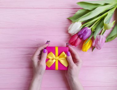 women's hands hold a gift box, a bouquet of tulips on a pink wooden background
