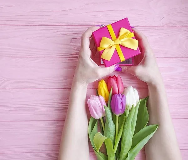 women's hands hold a gift box, a bouquet of tulips on a pink wooden background