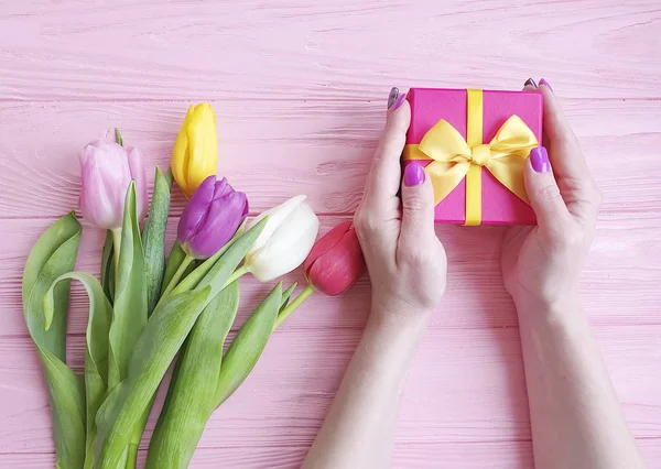 women's hands hold a gift box, a bouquet of tulips on a pink wooden background