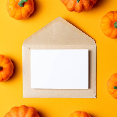 Üzerinde beyaz bir kart olan kahverengi Kraft zarfı, parlak turuncu arka planda küçük dekoratif balkabaklarıyla çevrili..