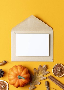 İçinde beyaz bir kart olan kahverengi Kraft zarfı, küçük balkabakları, kurutulmuş portakal dilimleri, tarçın çubukları ve parlak sarı arka planda meşe palamutları ile çevrili..