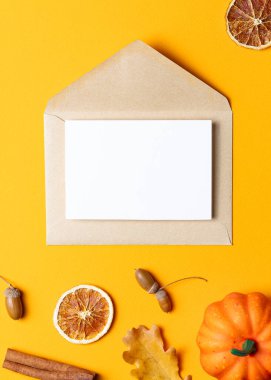 İçinde beyaz bir kart olan kahverengi Kraft zarfı, küçük balkabakları, kurutulmuş portakal dilimleri, tarçın çubukları ve parlak sarı arka planda meşe palamutları ile çevrili..