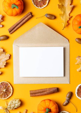 İçinde beyaz bir kart olan kahverengi Kraft zarfı, küçük balkabakları, kurutulmuş portakal dilimleri, tarçın çubukları ve parlak sarı arka planda meşe palamutları ile çevrili..