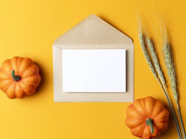 Üzerinde beyaz bir kart olan kahverengi Kraft zarfı, parlak turuncu arka planda küçük dekoratif balkabaklarıyla çevrili..