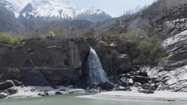 A breathtaking view of a waterfall cascading down a rocky cliff in the snowy mountains of Pakistan, with a small building and cable car system adding to the serene landscape.