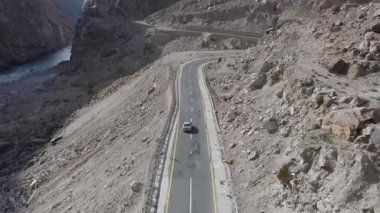 Aerial view of a winding road through the mountainous terrain of Pakistan, with a car navigating the serpentine path alongside a flowing river.