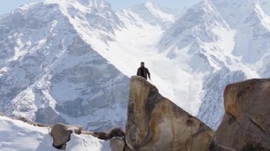 A solitary figure stands atop a rugged rock formation against the backdrop of towering snow-covered mountains in Pakistan.