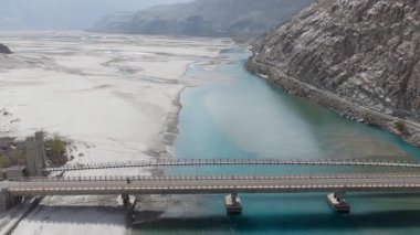 Aerial view of a turquoise river flowing through a snowy landscape in Pakistan, highlighting a solitary motorcycle crossing a bridge.