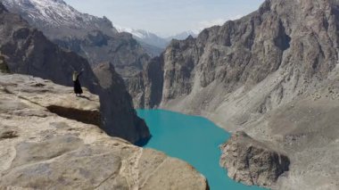 Aerial view of a breathtaking mountain valley in Pakistan, featuring a stunning turquoise lake, rugged peaks and a woman atop the cliff.