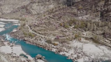 Pakistan 'da görkemli dağlar ve canlı bir nehirle çevrili sakin bir köyün havadan görünüşü geleneksel mimari ve yemyeşil alanları vurguluyor..