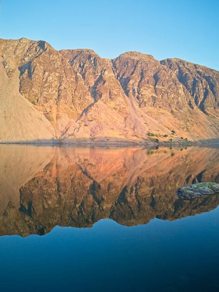 Berrak mavi gökyüzünün altındaki sakin göle mükemmel bir şekilde yansıyan bir dağ sırası. Çarpıcı bir doğal manzara..