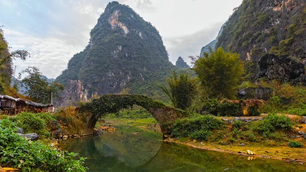 Sakin bir nehrin üzerindeki yeşil kemerlerle kaplı antik bir taş köprü. Görkemli dağlar ve yemyeşil bitki örtüsü, cennet manzarasının etrafını sarar..