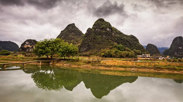 Bereketli bir yeşillik ve bulutlu bir gökyüzü tarafından çerçevelenmiş sakin bir nehirde yansıtılan, yüksek dağlara ev sahipliği yapar..