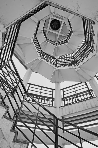 Siyah ve beyaz bir fotoğraf, geometrik bir kulenin içindeki sarmal bir merdivenin yukarı doğru görüntüsünü yakalar ve mimari detayları vurgular..