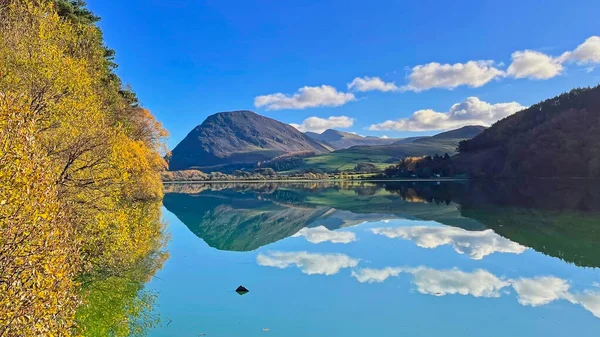 Sakin bir gölün çarpıcı manzarası bir dağı, bulutları ve mavi gökyüzünü yansıtıyor. Altın ağaçlar sonbaharda kıyıya dizilir..