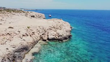 Kıbrıs 'ın kayalık kıyı şeridinde seyreden bir teknenin hava görüntüsü. Güzel kıyı manzarasında macera teknesi gezisi konsepti. Mavi Akdeniz suyu ve bir yelkenli.