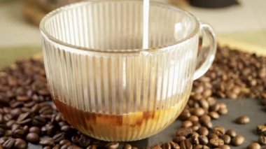 Süt, kahve çekirdeğinin arka planında kahve fincanına dökülür. Sıcak içecekten buhar çıkıyor. Yakın plan. Yüksek kaliteli FullHD görüntüler