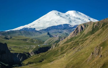 Elbrus görünümü - en yüksek tepe Rusya'nın ve Avrupa peyzaj. Beyaz kar dağ veya volkan Elbrus yeşil alanları, mavi gökyüzü arka plan ile inanılmaz bakış açısı