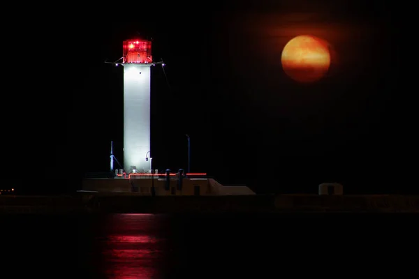 Vorontsov Farol noite contra a ascensão da grande lua vermelha. Velho farol de lanterna farol branco no porto do Mar Negro de Odessa, Ucrânia. Exposição longa . — Fotografia de Stock