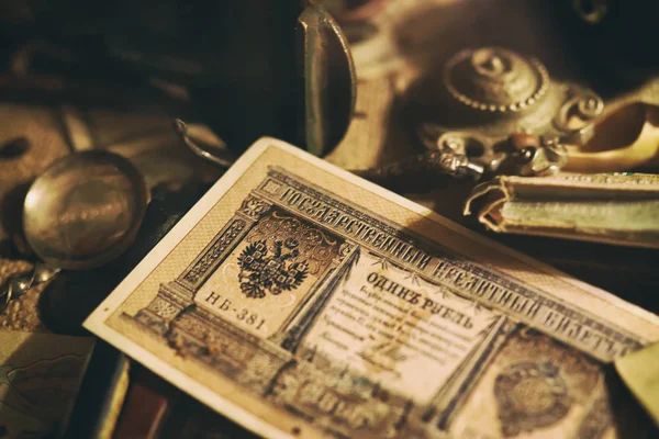 桌上摆着不同的古董：青铜首饰、俄罗斯帝国的纸币和硬币、一箱眼镜、银器。古董收藏的复古背景。特写选定焦点 — 图库照片