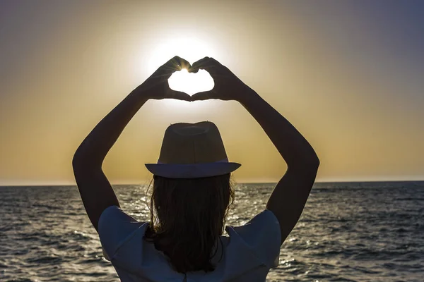 Silhouette of a young woman creating the shape of a heart with her hands with the sun nearly inside