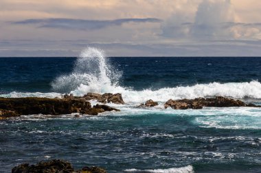 Hawaii'nin Büyük Adası'ndaki Black Sand Beach'te (Punaluuu) koyu volkanik kayaya düşen dalga. Beyaz sprey havada yükselir; Mavi Pasifik Okyanusu, gökyüzü ve bulutlar arka planda. 