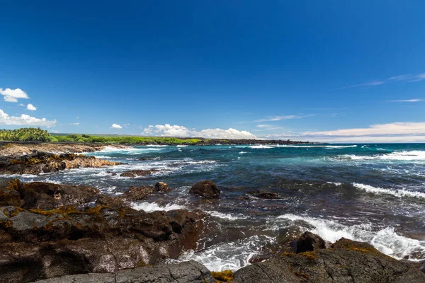 Ocean view at Black Sand Beach on Hawaii\'s Big Island. Blue pacific ocean with waves, rough volcanic rock and brown vegetation on shore. Coastline with green plants in the distance.