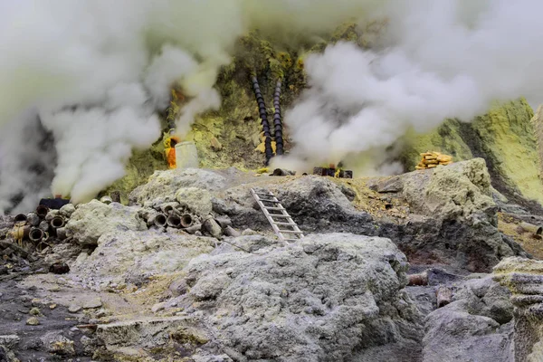 Sulfur mining operation in Mount Ijen crater, East Java, Indonesia. White volcanic gas rising from fissues in the ground. Pipes in background to divert the smoke for condensation. Yellow sulfur scattered across the ground.