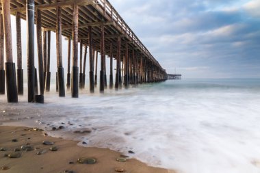 Ventura Pier, Ventura, California. Early light from sunrise visible from left.  sand and rocks in foreground; smooth incoming wave reaching shore; ocean, cloudy sky beyond.  clipart