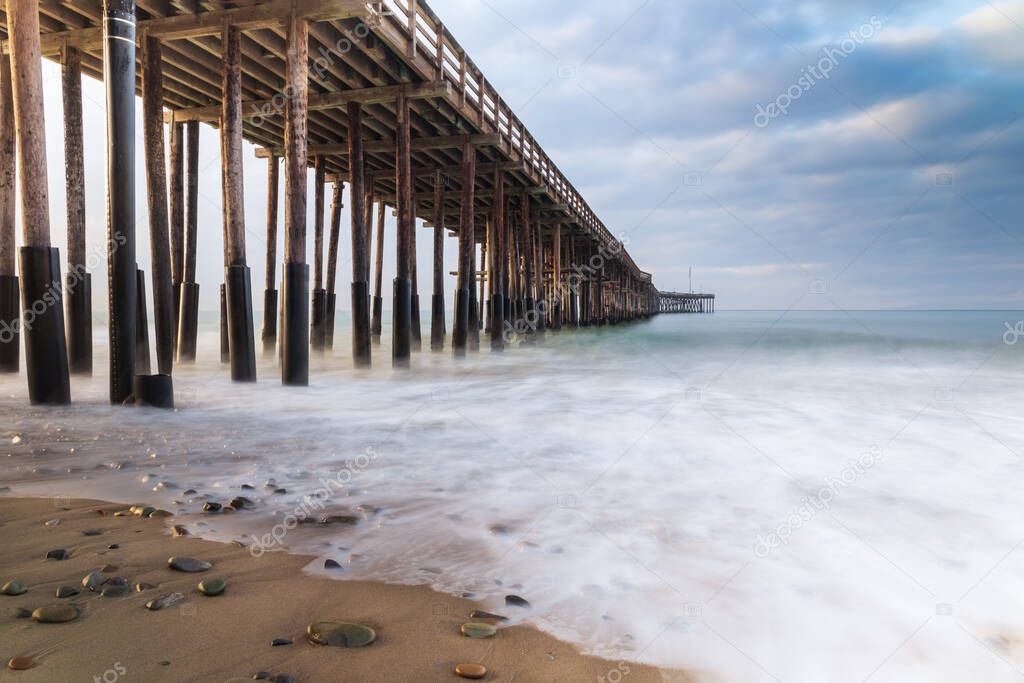 Ventura Pier, Ventura, California. Early light from sunrise visible from left.  sand and rocks in foreground; smooth incoming wave reaching shore; ocean, cloudy sky beyond. 