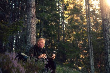 Ağaçlar ve güneş ışığıyla çevrili akıllı bir telefonla ormanda oturan bir adam..