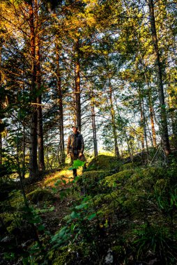 İsveç 'te bir orman yolunda duran sırt çantalı bir gezginin dikey görüntüsü. Vücudu kameraya doğru ama sola bakıyor. Yosun kaplı kaya, çalılar ve yüksek ağaçlar doğal bir huzur ortamı yaratır..