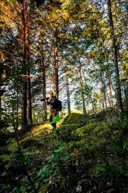 Ormanda elinde uzun bir telefoto kamera lensi olan bir adamın dikey görüntüsü çalılar ve arka planda uzun ağaçlar olan yosun kaplı kayalar arasında dar bir yolda duruyor. Çerçevede küçük, arkasında güneş var.